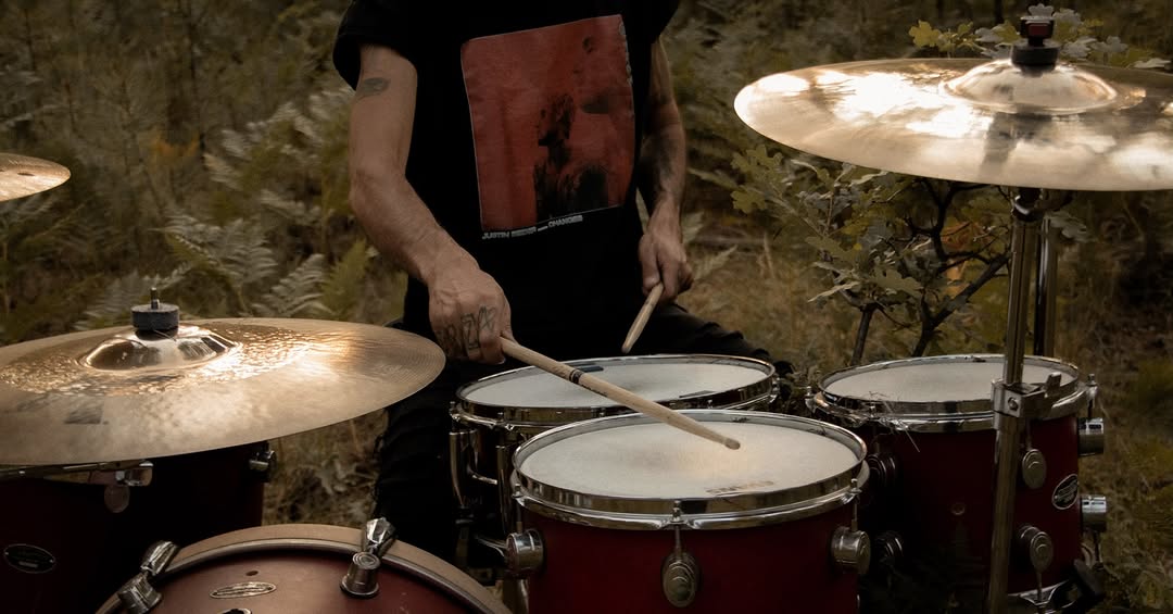 Close, wide frame of a drummer mid-hit on a deep red kit surrounded by ferns in the forest