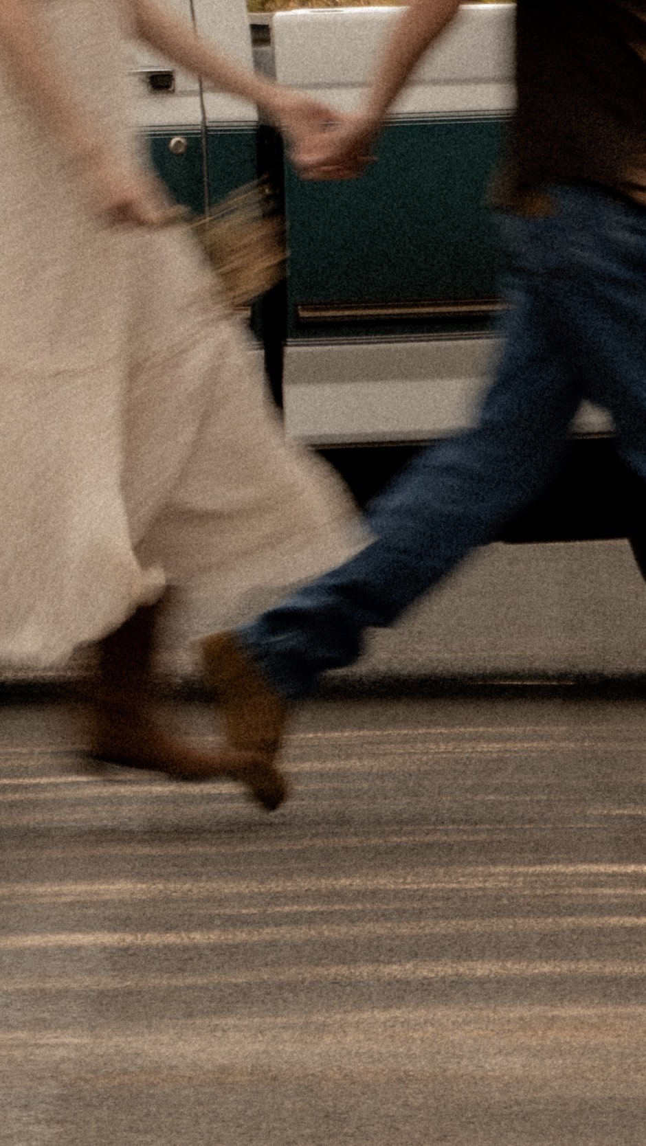 A just-married couple mid-stride, hands clasped, dress and boots in motion beside their getaway truck